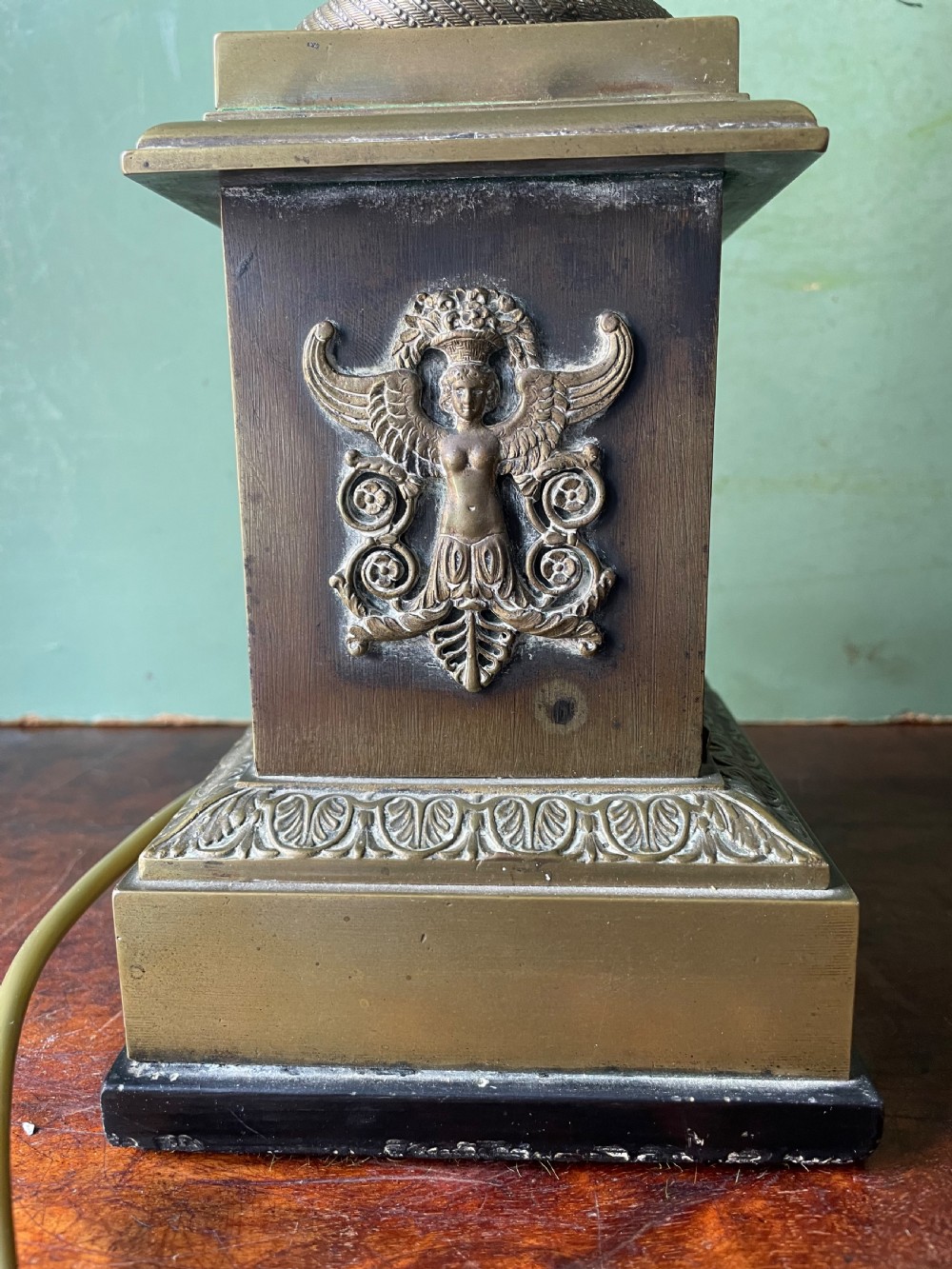 An early nineteenth-century patinated brass table lamp featuring a Corinthian capital, embossed shaft, and winged relief figure on a square stepped base. View XI