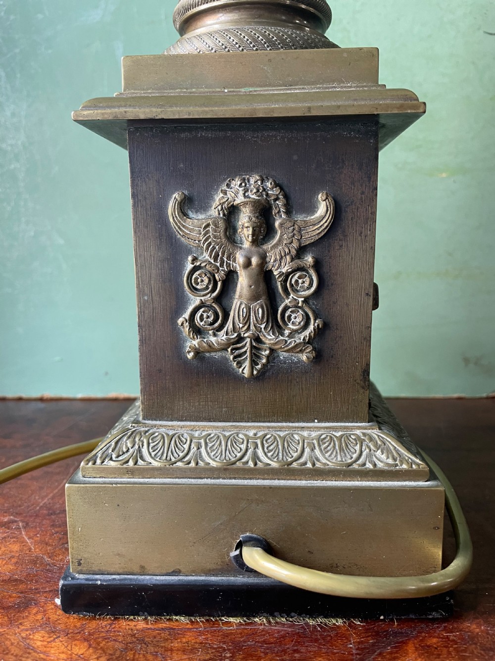 An early nineteenth-century patinated brass table lamp featuring a Corinthian capital, embossed shaft, and winged relief figure on a square stepped base. View XIII