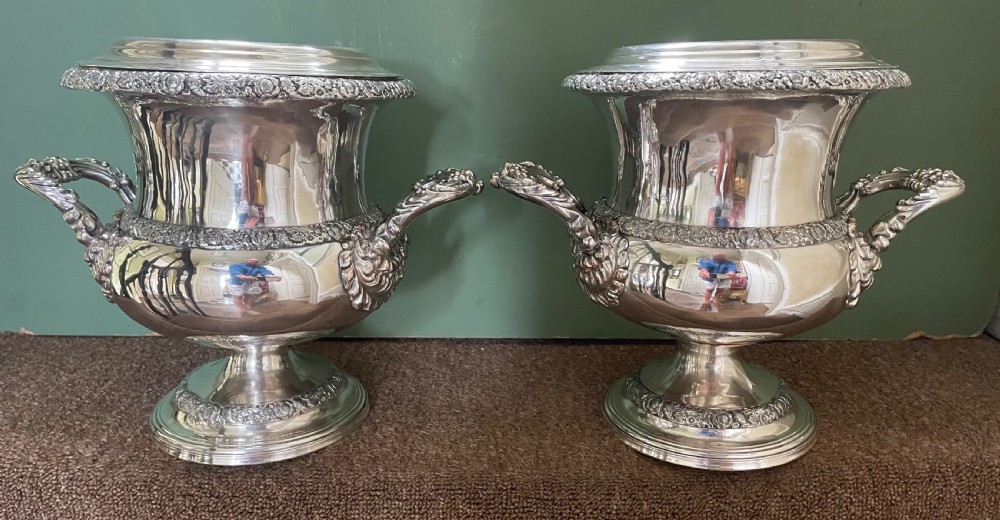 A pair of polished silver-plated campana-form wine coolers featuring ornate foliate handles, intricate relief borders, and pedestal bases.