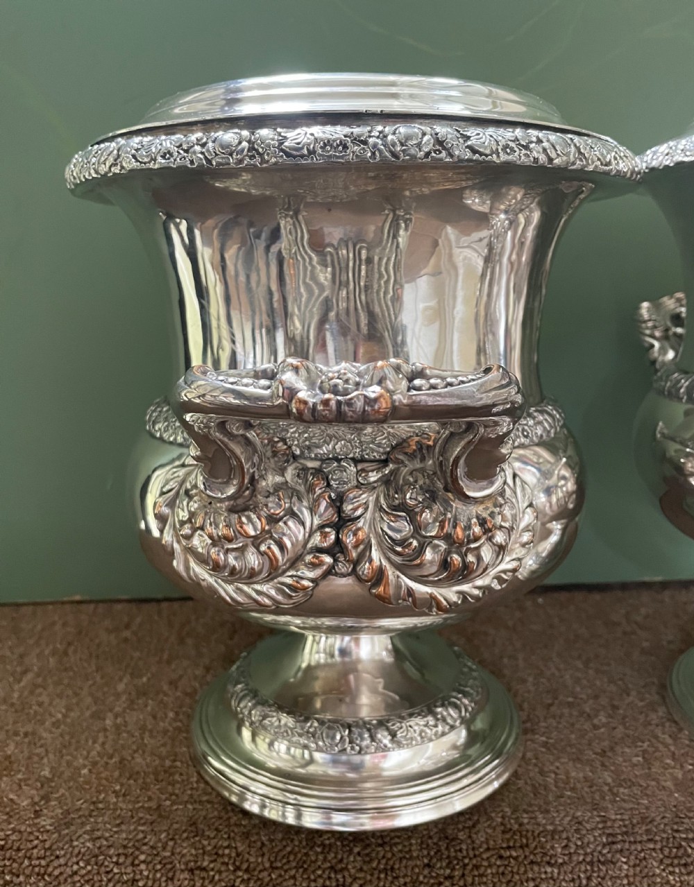 A pair of polished silver-plated campana-form wine coolers featuring ornate foliate handles, intricate relief borders, and pedestal bases. View VI