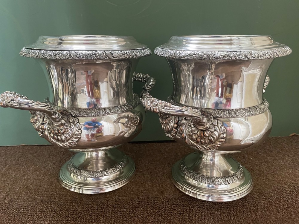A pair of polished silver-plated campana-form wine coolers featuring ornate foliate handles, intricate relief borders, and pedestal bases. View IX