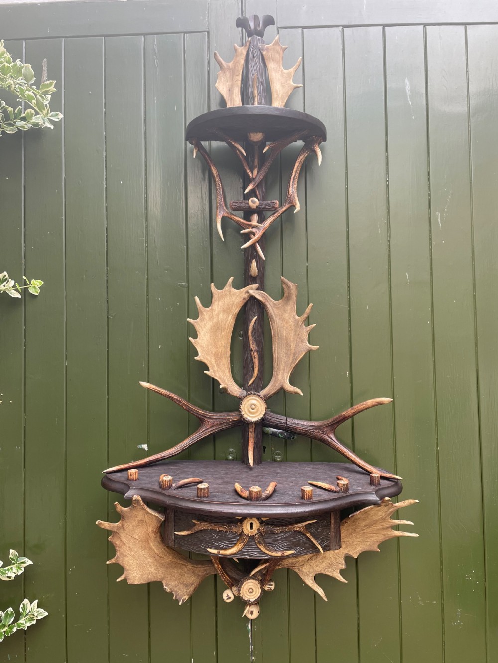 A nineteenth-century Black Forest carved wood wall rack with natural deer antlers, small shelves, and coat hooks against a green background.