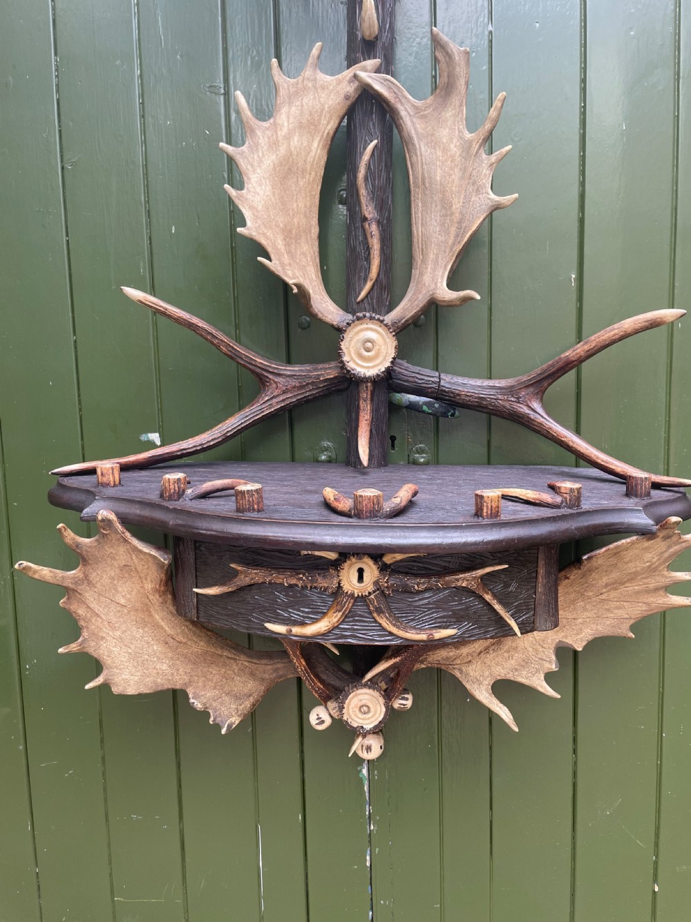 A nineteenth-century Black Forest carved wood wall rack with natural deer antlers, small shelves, and coat hooks against a green background. View II