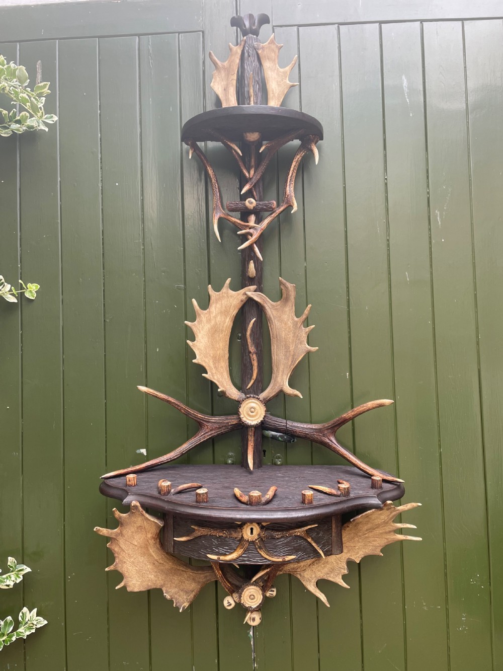 A nineteenth-century Black Forest carved wood wall rack with natural deer antlers, small shelves, and coat hooks against a green background. View V