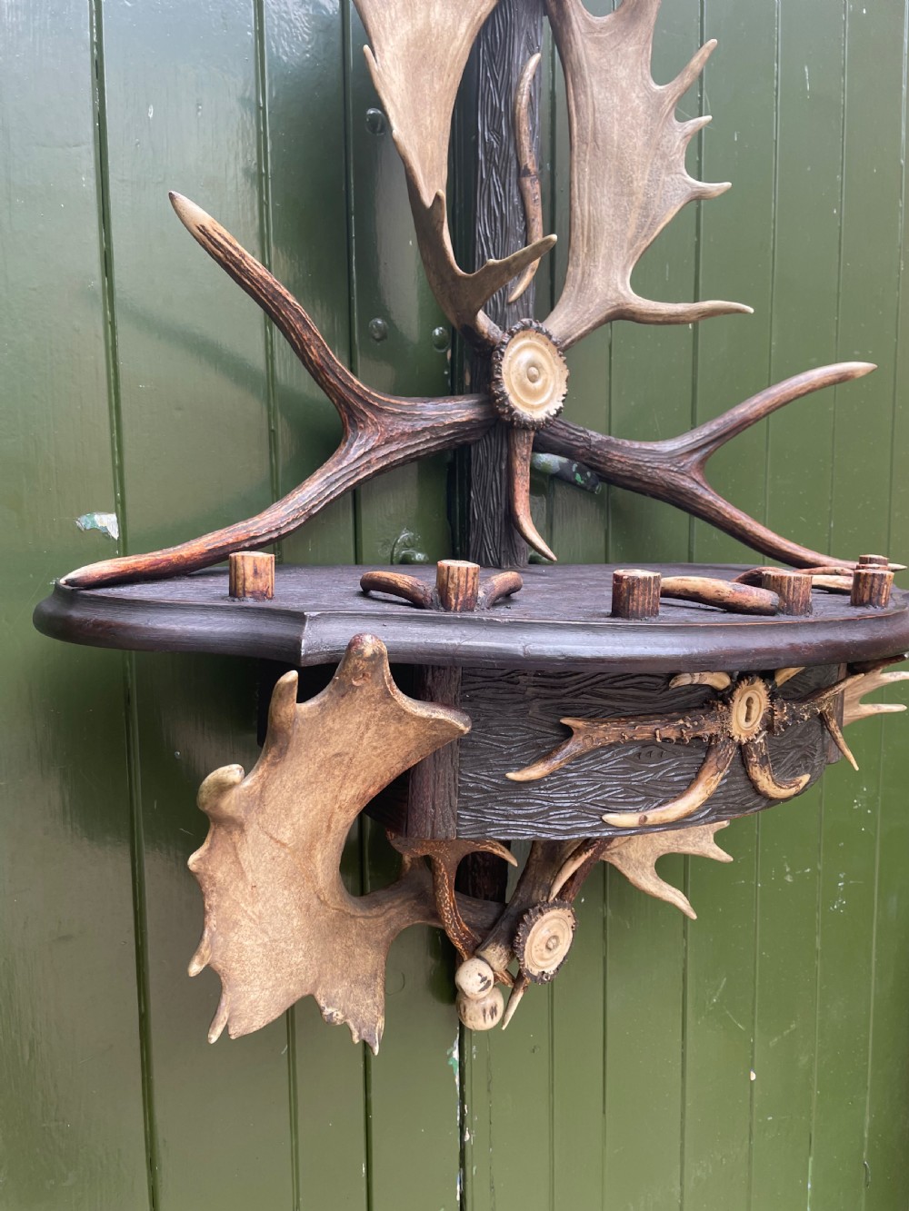 A nineteenth-century Black Forest carved wood wall rack with natural deer antlers, small shelves, and coat hooks against a green background. View VI