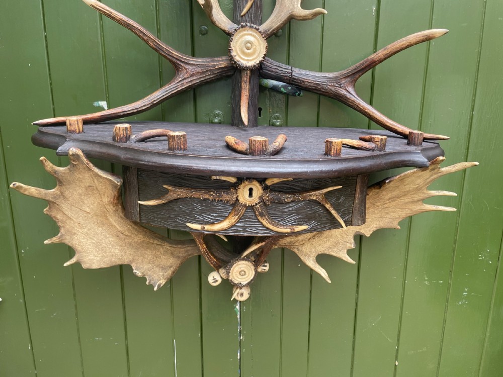 A nineteenth-century Black Forest carved wood wall rack with natural deer antlers, small shelves, and coat hooks against a green background. View IX
