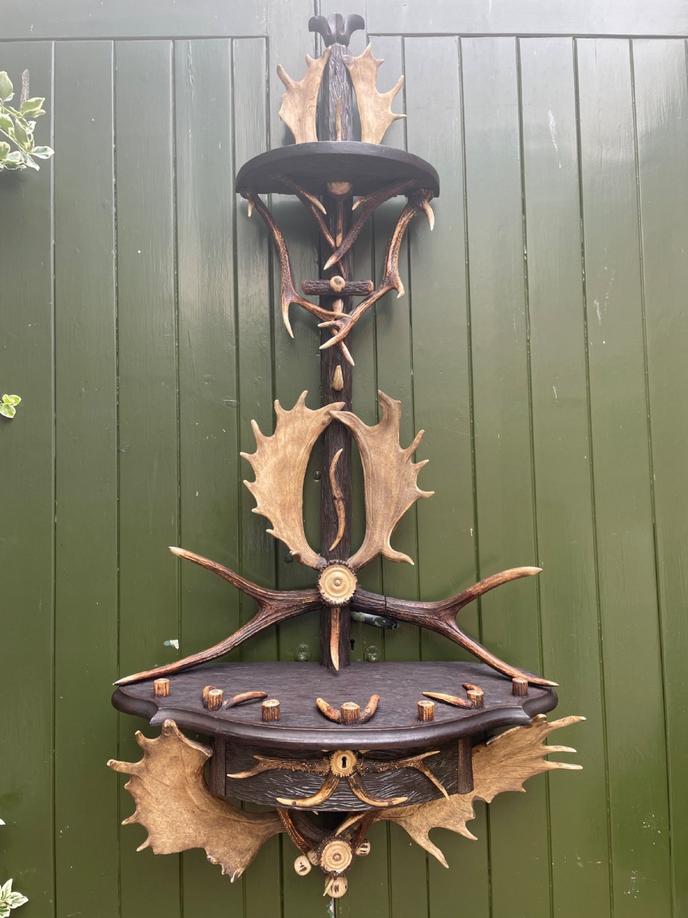 A nineteenth-century Black Forest carved wood wall rack with natural deer antlers, small shelves, and coat hooks against a green background. View XII