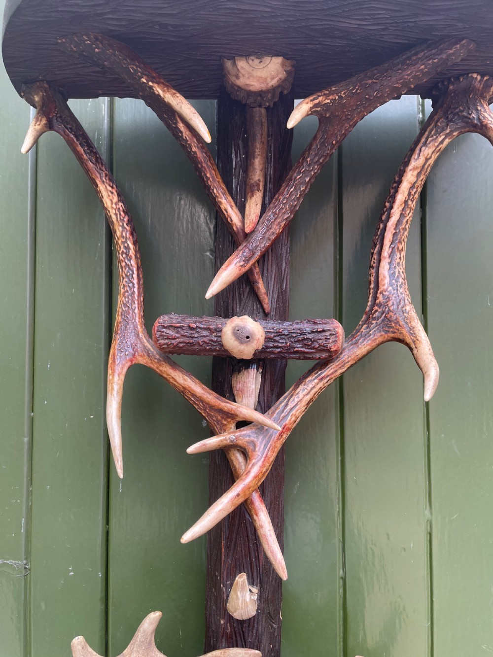 A nineteenth-century Black Forest carved wood wall rack with natural deer antlers, small shelves, and coat hooks against a green background. View XIII