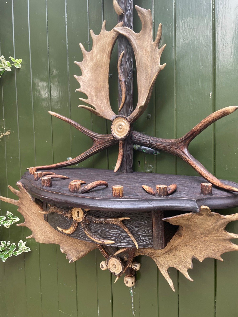 A nineteenth-century Black Forest carved wood wall rack with natural deer antlers, small shelves, and coat hooks against a green background. View XVIII