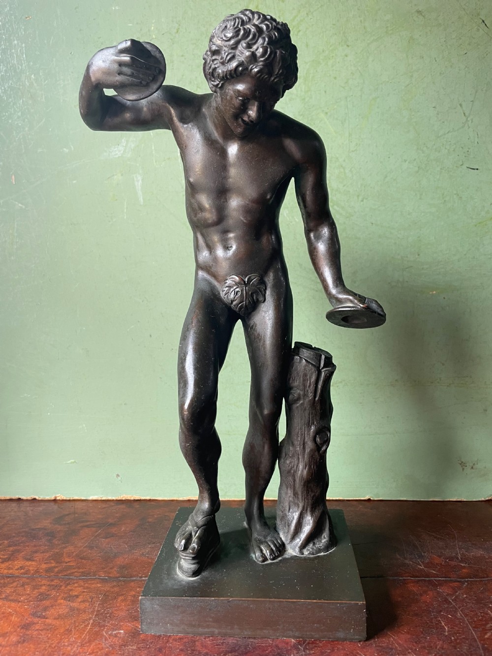 A dark patinated bronze sculpture depicting the classical figure of the Dancing Faun holding cymbals, mounted on a rectangular metal plinth.