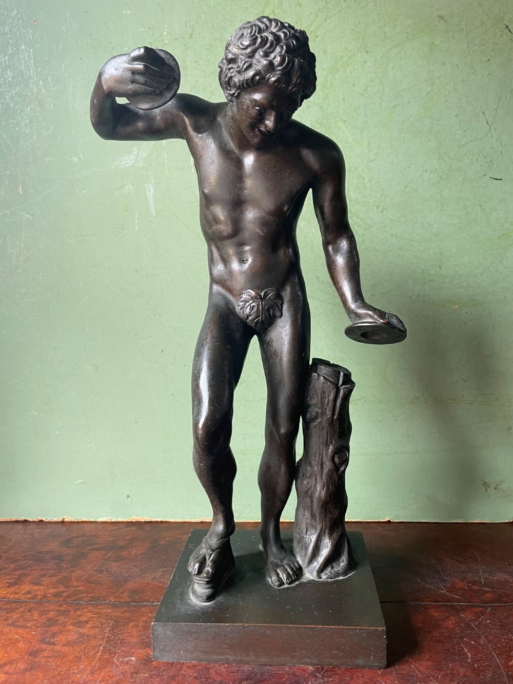 A dark patinated bronze sculpture depicting the classical figure of the Dancing Faun holding cymbals, mounted on a rectangular metal plinth. View II
