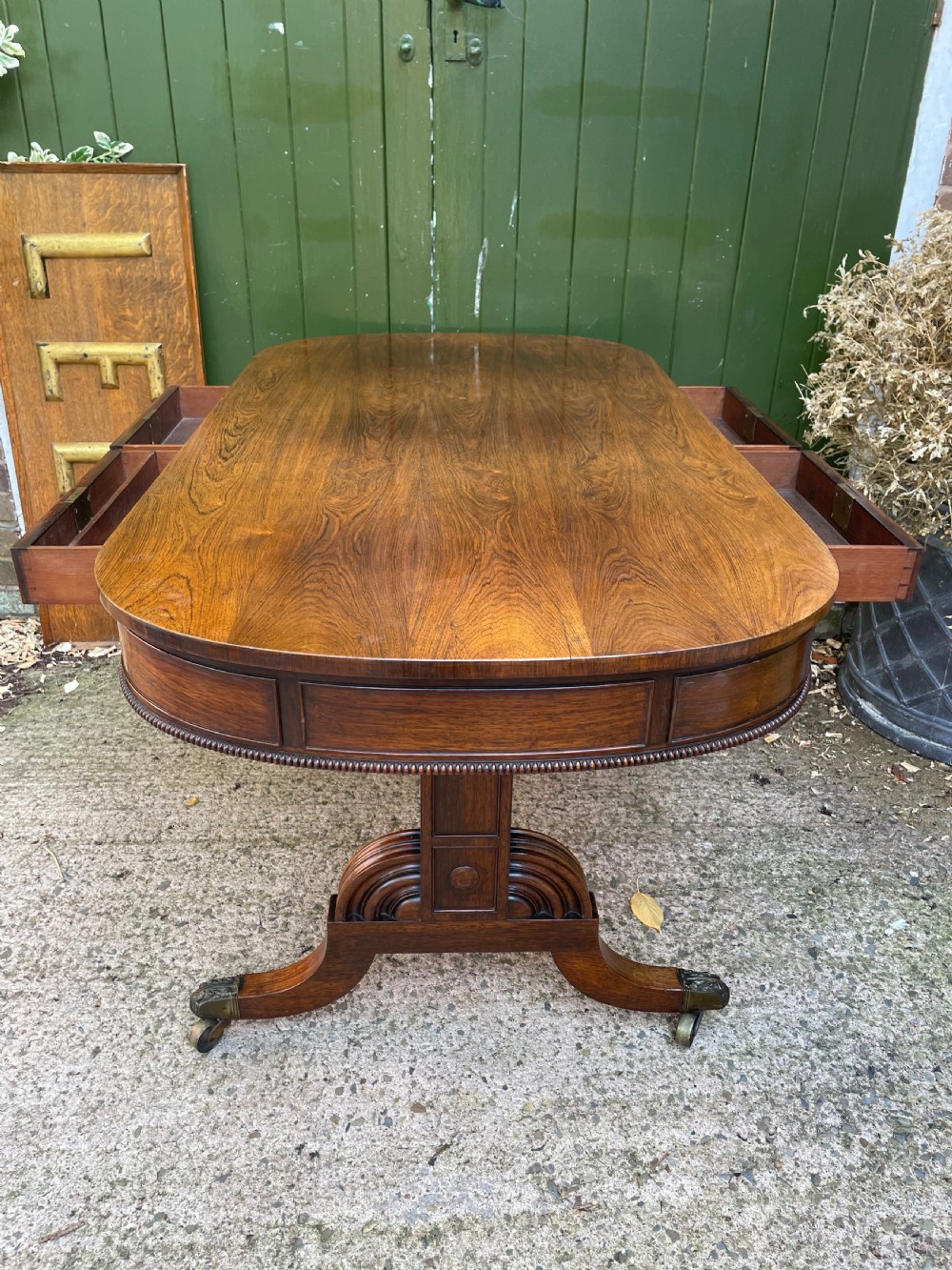 An early nineteenth-century George IV rosewood library table featuring two frieze drawers, a turned horizontal stretcher, and trestle legs on brass castors. View X