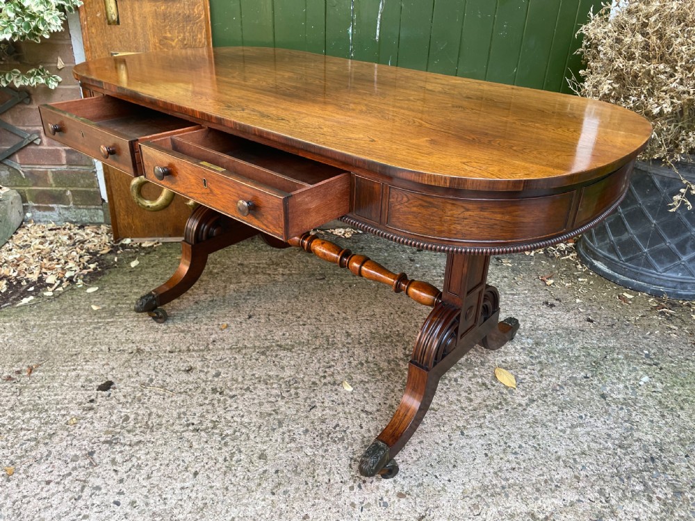 An early nineteenth-century George IV rosewood library table featuring two frieze drawers, a turned horizontal stretcher, and trestle legs on brass castors. View XIII