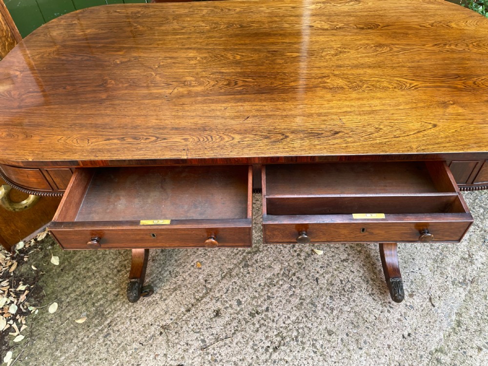 An early nineteenth-century George IV rosewood library table featuring two frieze drawers, a turned horizontal stretcher, and trestle legs on brass castors. View XIV