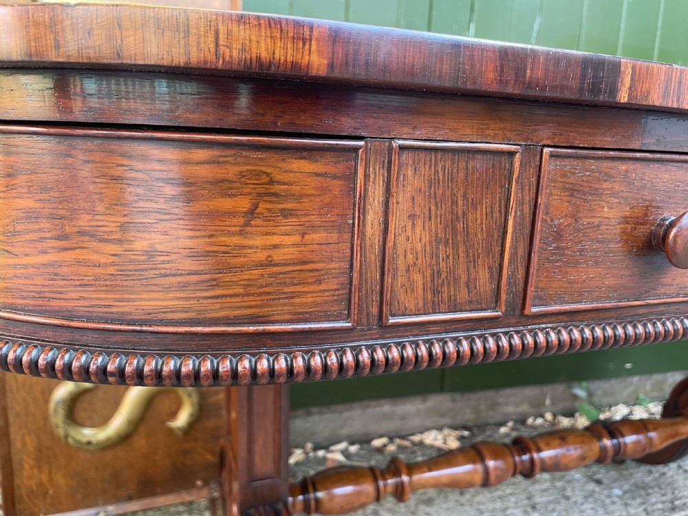 An early nineteenth-century George IV rosewood library table featuring two frieze drawers, a turned horizontal stretcher, and trestle legs on brass castors. View XVII