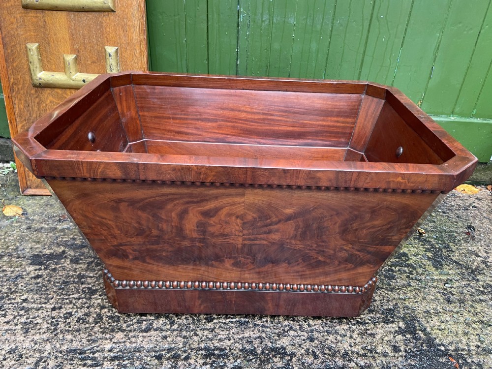 A George IV open mahogany wine cooler of classical sarcophagus form featuring flame mahogany veneer panels and carved bead moulding. View II