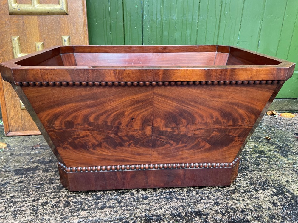 A George IV open mahogany wine cooler of classical sarcophagus form featuring flame mahogany veneer panels and carved bead moulding. View X
