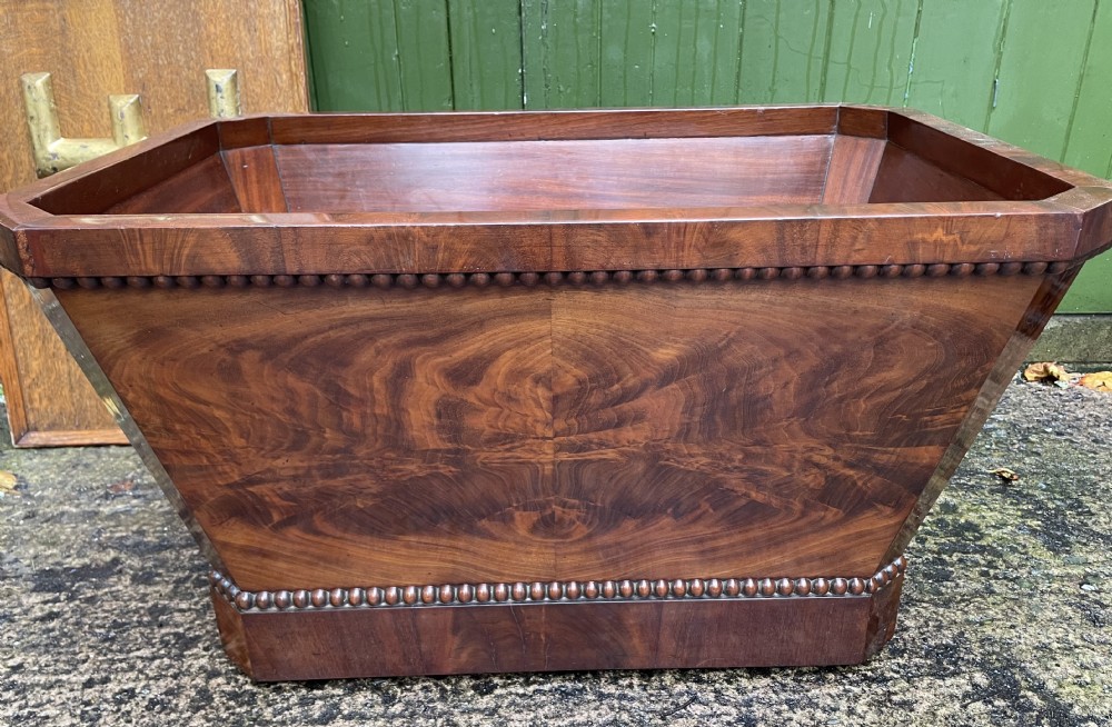 A George IV open mahogany wine cooler of classical sarcophagus form featuring flame mahogany veneer panels and carved bead moulding.