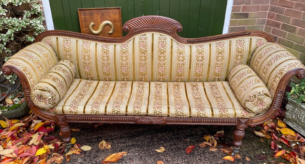 An early nineteenth-century Regency mahogany sofa featuring double scroll-ended arms, a carved crest rail, striped gold velvet upholstery, matching bolster cushions, and turned legs on brass castors. View VI