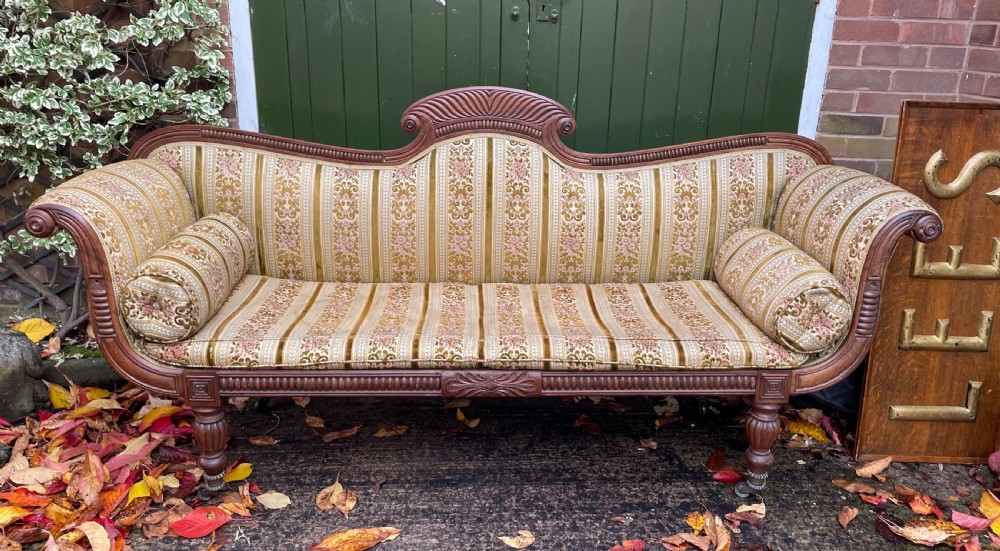 An early nineteenth-century Regency mahogany sofa featuring double scroll-ended arms, a carved crest rail, striped gold velvet upholstery, matching bolster cushions, and turned legs on brass castors. View XI