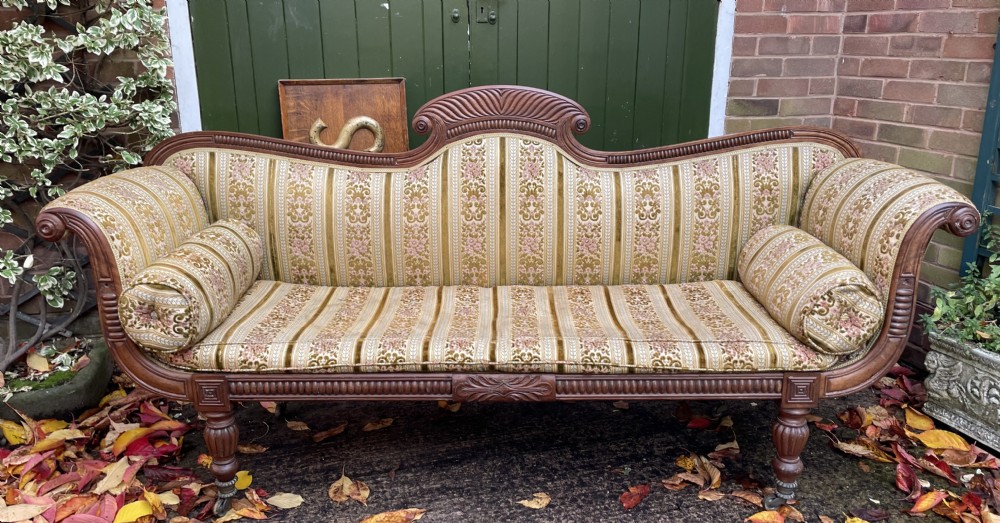 An early nineteenth-century Regency mahogany sofa featuring double scroll-ended arms, a carved crest rail, striped gold velvet upholstery, matching bolster cushions, and turned legs on brass castors. View XIII