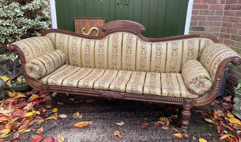 An early nineteenth-century Regency mahogany sofa featuring double scroll-ended arms, a carved crest rail, striped gold velvet upholstery, matching bolster cushions, and turned legs on brass castors. View II