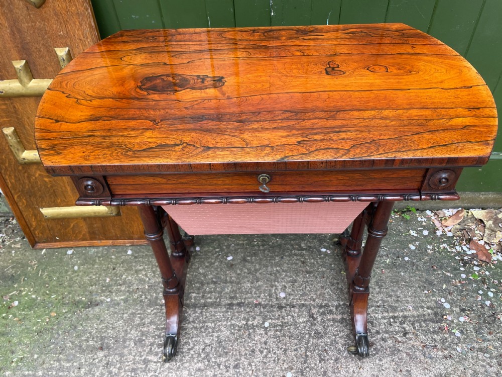 An early 19th-century George IV figured rosewood work table featuring a smooth polished top, single drawer, fabric work basket, turned twin-column supports, and brass castors. View II