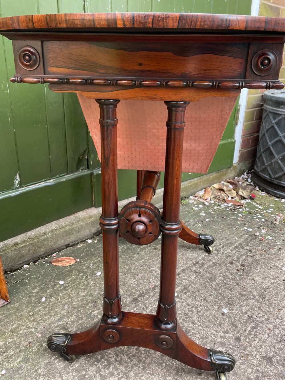 An early 19th-century George IV figured rosewood work table featuring a smooth polished top, single drawer, fabric work basket, turned twin-column supports, and brass castors. View III