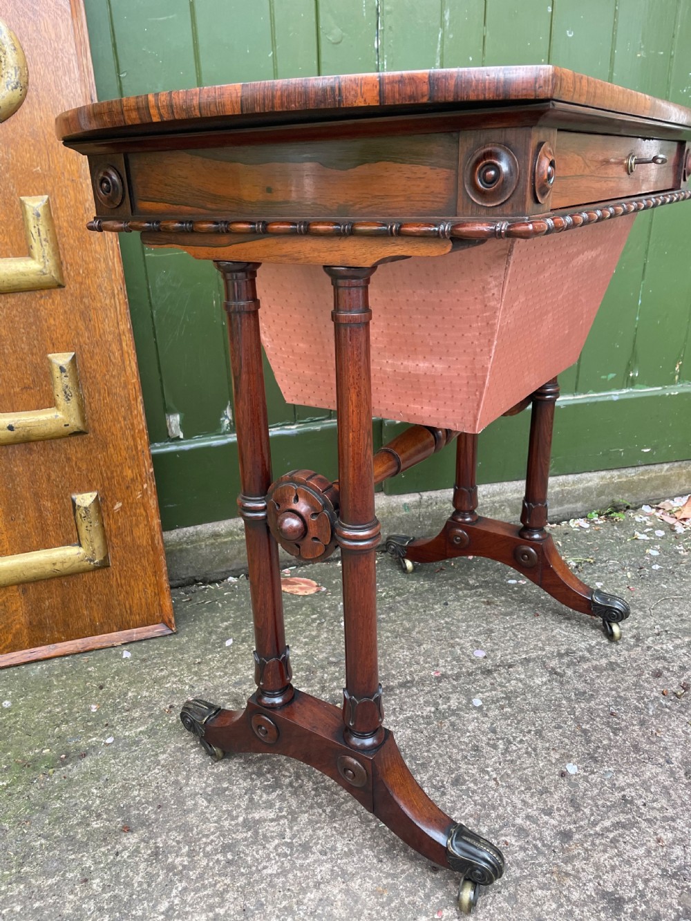 An early 19th-century George IV figured rosewood work table featuring a smooth polished top, single drawer, fabric work basket, turned twin-column supports, and brass castors. View V