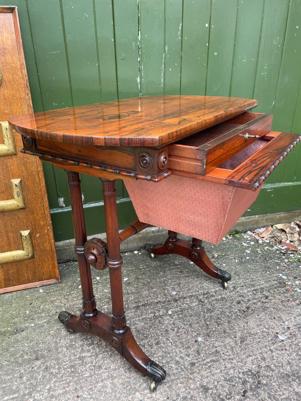 An early 19th-century George IV figured rosewood work table featuring a smooth polished top, single drawer, fabric work basket, turned twin-column supports, and brass castors. View VI