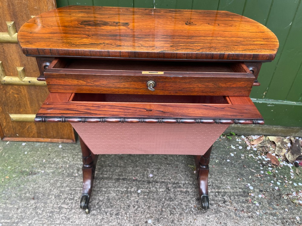 An early 19th-century George IV figured rosewood work table featuring a smooth polished top, single drawer, fabric work basket, turned twin-column supports, and brass castors. View VII