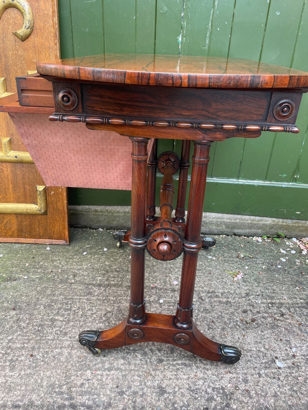 An early 19th-century George IV figured rosewood work table featuring a smooth polished top, single drawer, fabric work basket, turned twin-column supports, and brass castors. View X
