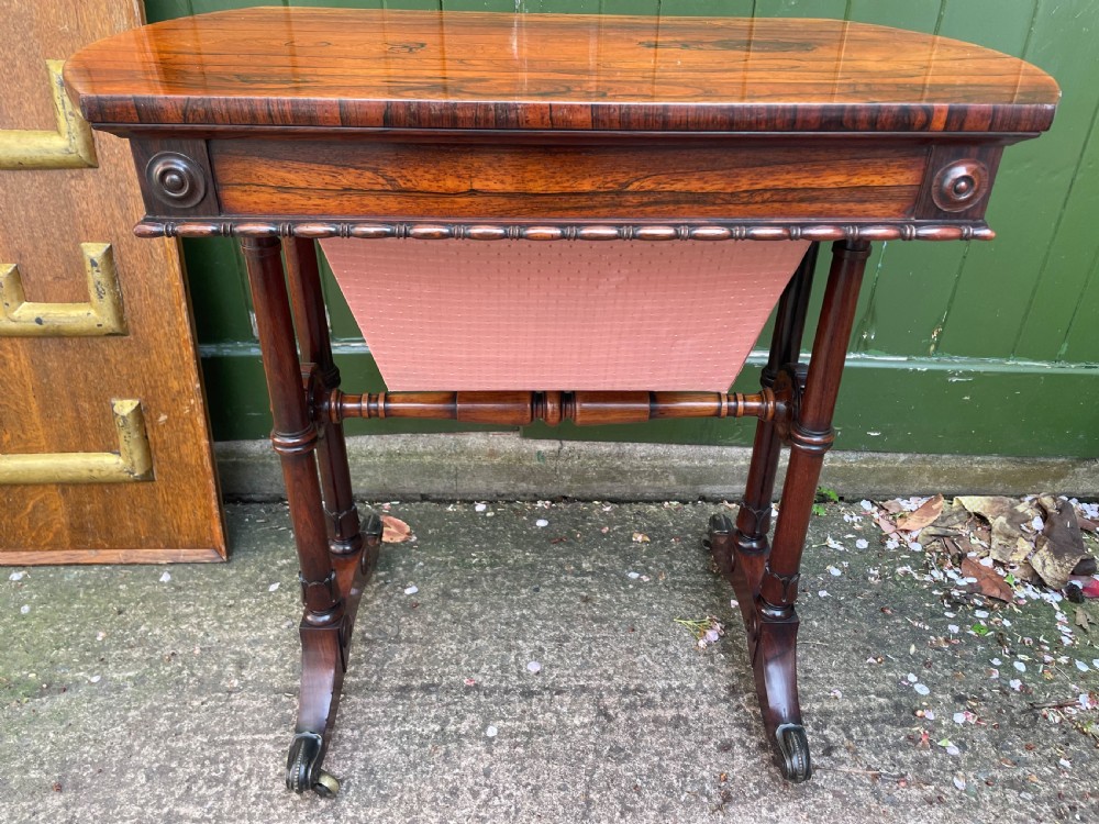 An early 19th-century George IV figured rosewood work table featuring a smooth polished top, single drawer, fabric work basket, turned twin-column supports, and brass castors. View XI