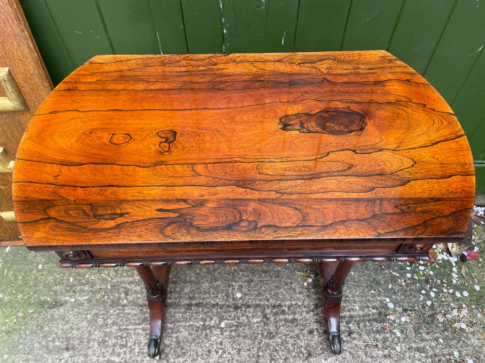An early 19th-century George IV figured rosewood work table featuring a smooth polished top, single drawer, fabric work basket, turned twin-column supports, and brass castors. View XIV