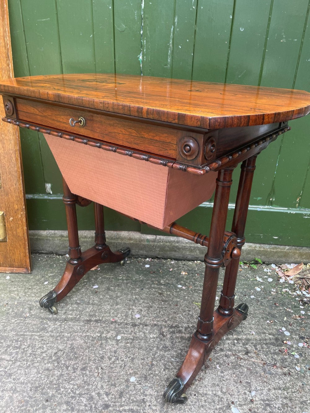 An early 19th-century George IV figured rosewood work table featuring a smooth polished top, single drawer, fabric work basket, turned twin-column supports, and brass castors. View XV