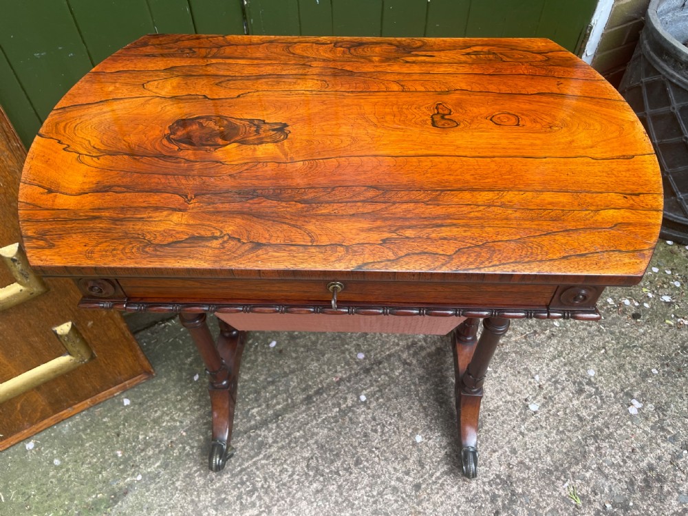 An early 19th-century George IV figured rosewood work table featuring a smooth polished top, single drawer, fabric work basket, turned twin-column supports, and brass castors. View XVII