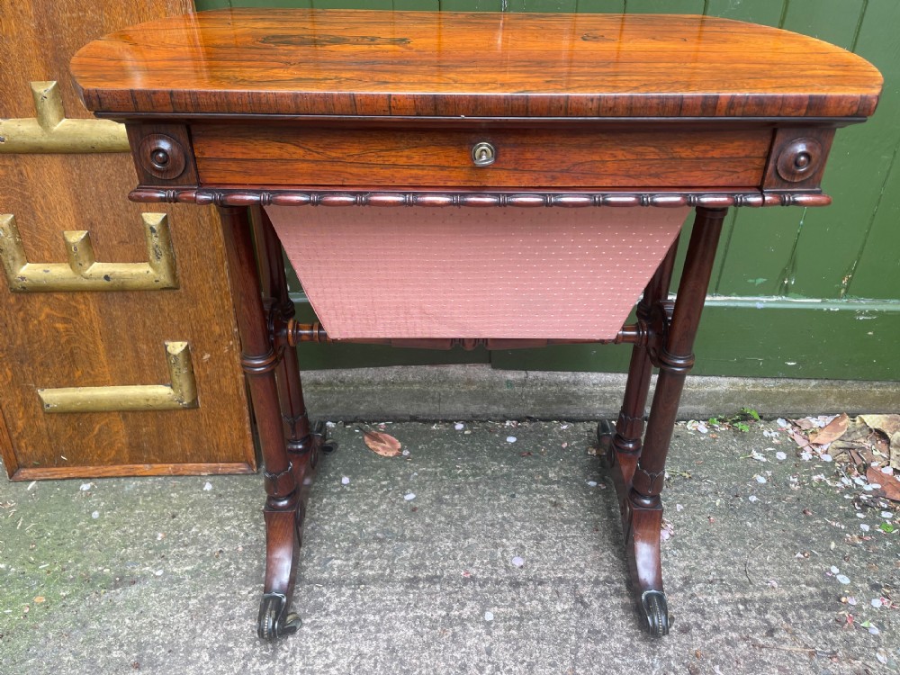 An early 19th-century George IV figured rosewood work table featuring a smooth polished top, single drawer, fabric work basket, turned twin-column supports, and brass castors. View XIX