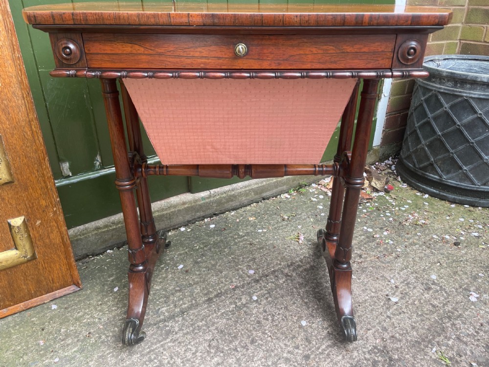 An early 19th-century George IV figured rosewood work table featuring a smooth polished top, single drawer, fabric work basket, turned twin-column supports, and brass castors. View XX
