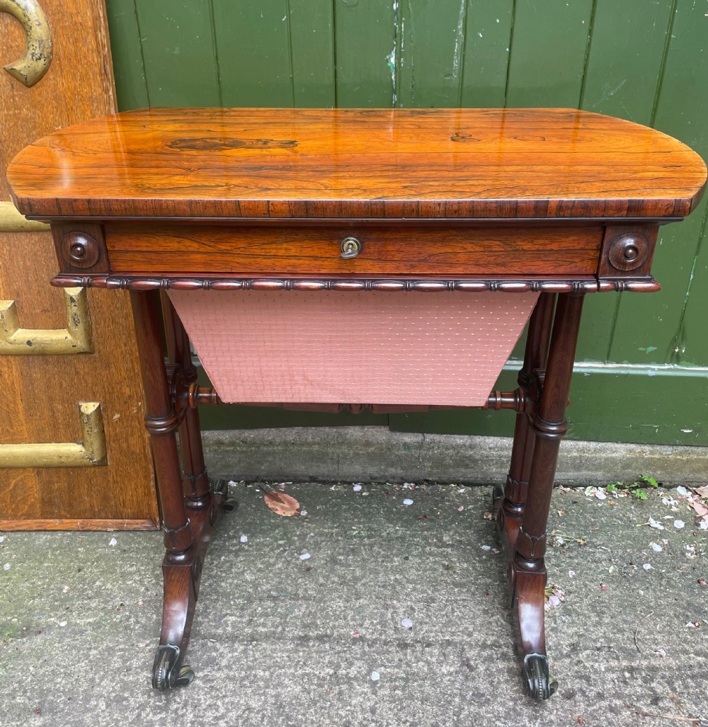 An early 19th-century George IV figured rosewood work table featuring a smooth polished top, single drawer, fabric work basket, turned twin-column supports, and brass castors. View XXI