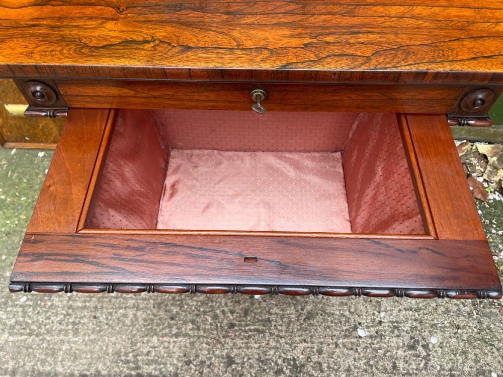 An early 19th-century George IV figured rosewood work table featuring a smooth polished top, single drawer, fabric work basket, turned twin-column supports, and brass castors. View VIII