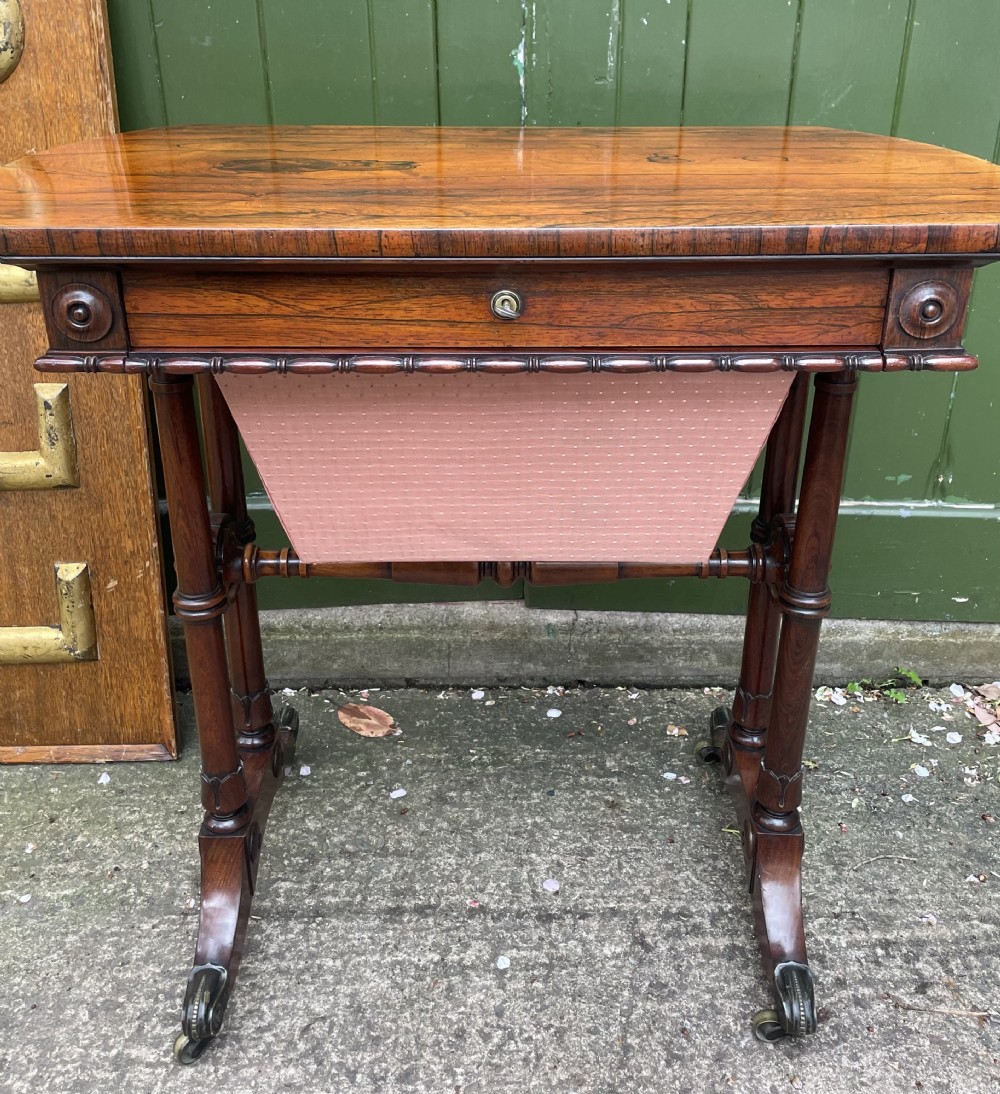 An early 19th-century George IV figured rosewood work table featuring a smooth polished top, single drawer, fabric work basket, turned twin-column supports, and brass castors. View XVI