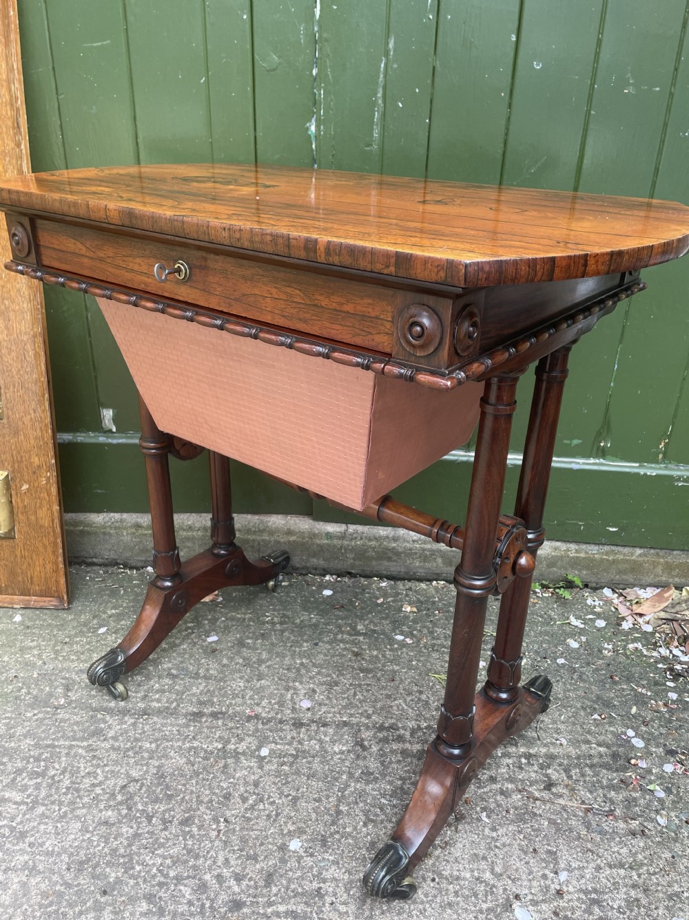 An early 19th-century George IV figured rosewood work table featuring a smooth polished top, single drawer, fabric work basket, turned twin-column supports, and brass castors. View XXVII