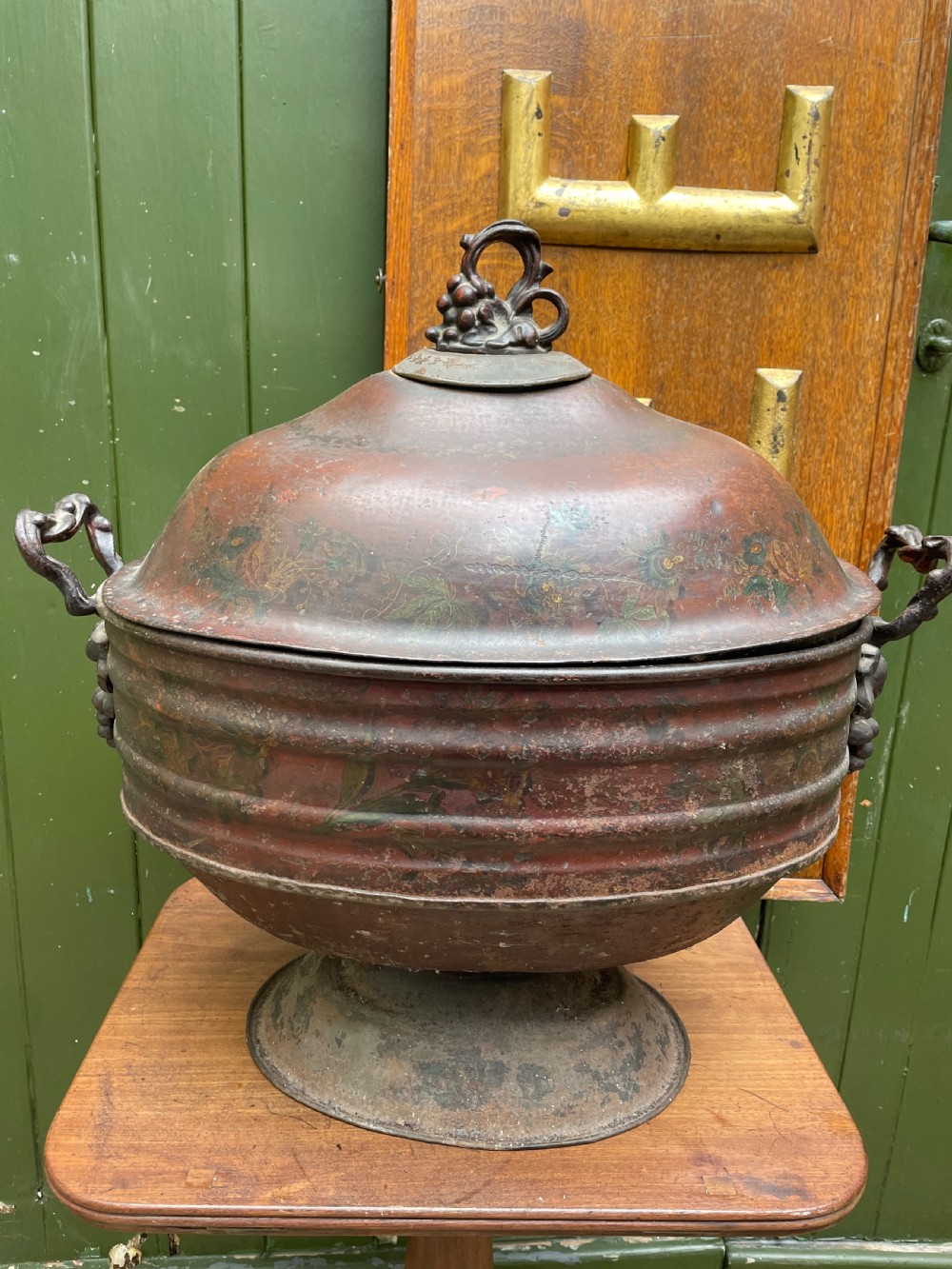 An early nineteenth-century round japanned tinware wine cooler with a domed lid, pedestal base, twin loop handles, and faded painted floral motifs. View XVI