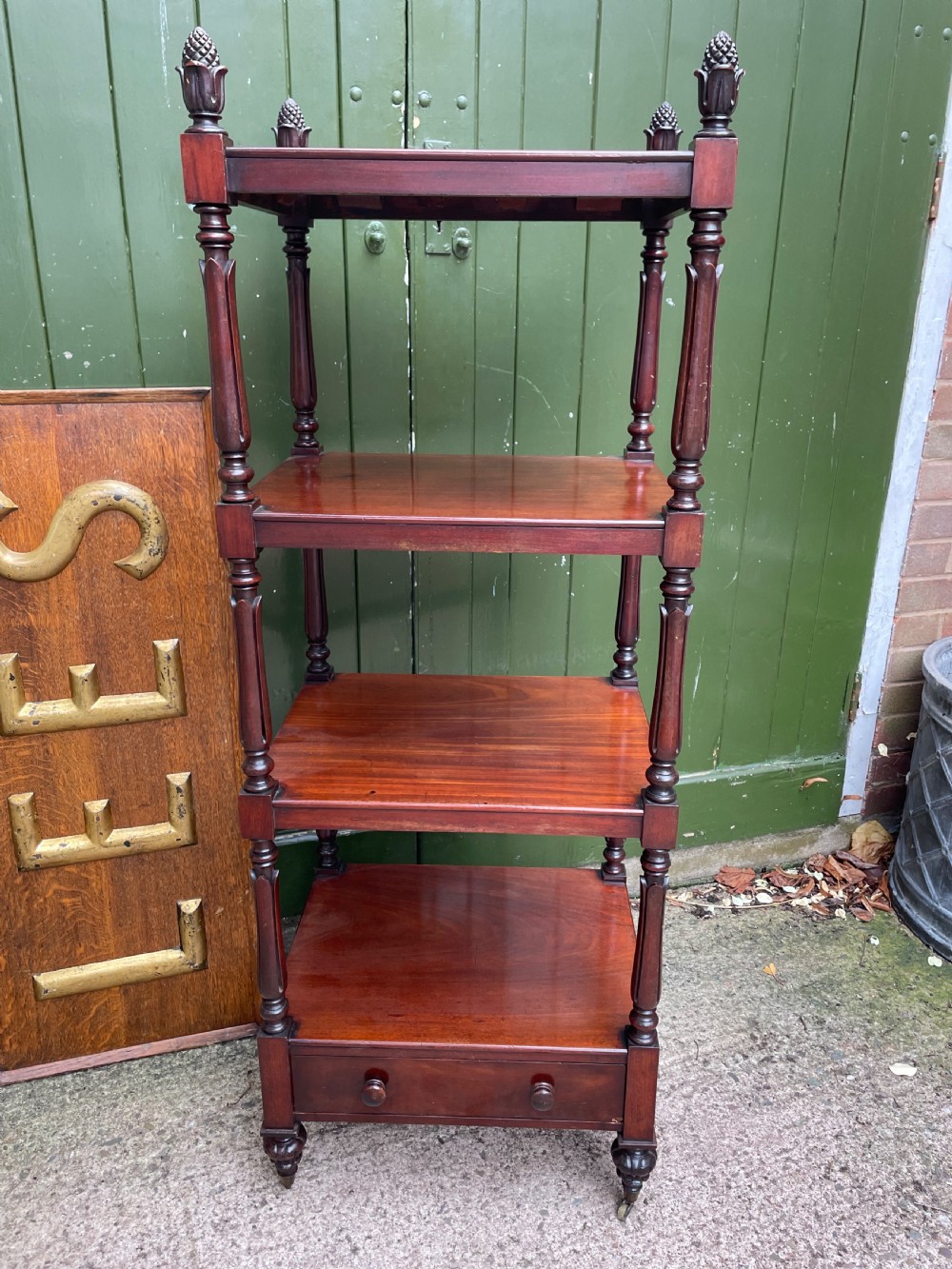 An early nineteenth-century George IV mahogany four-tier etagere featuring carved pinecone finials, turned supports, a single lower drawer, and brass castors. View V