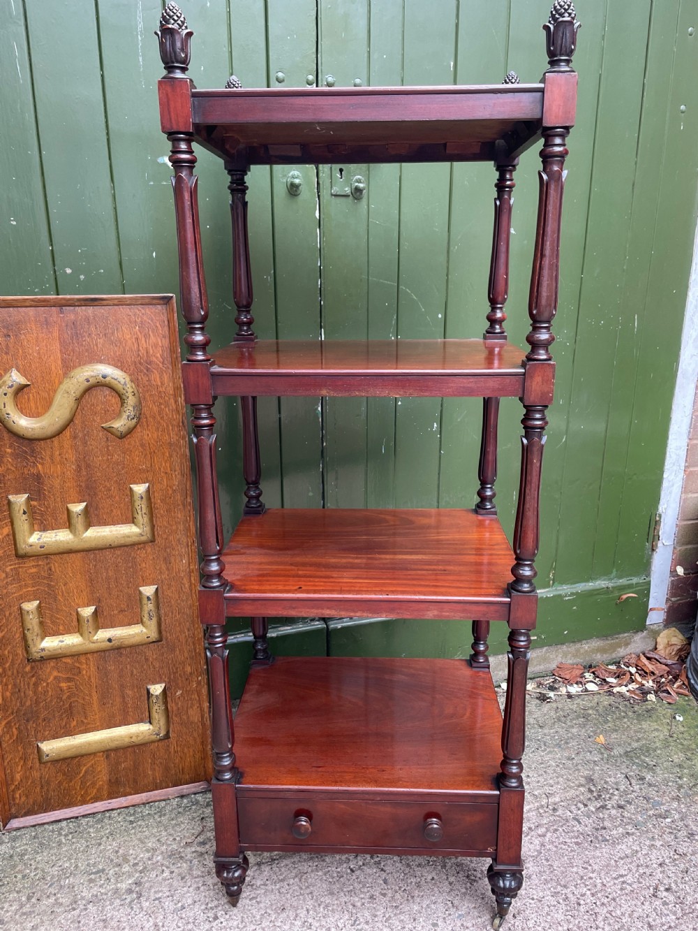 An early nineteenth-century George IV mahogany four-tier etagere featuring carved pinecone finials, turned supports, a single lower drawer, and brass castors. View X