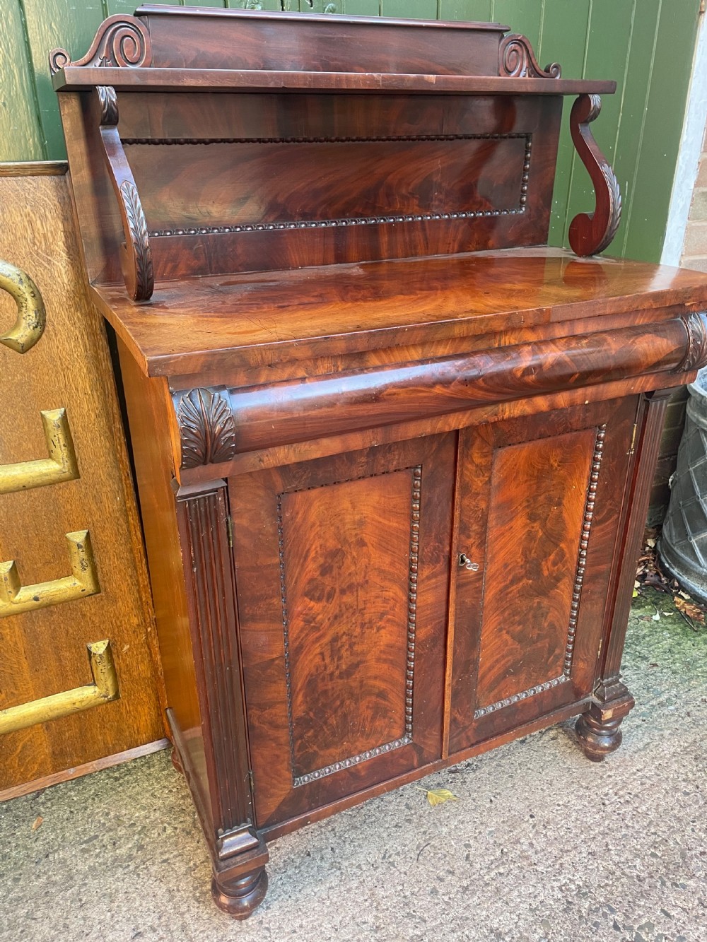An early nineteenth-century George IV flame mahogany chiffonier featuring a raised shelf gallery with scroll supports above a two-door cabinet supported on turned bun feet.
