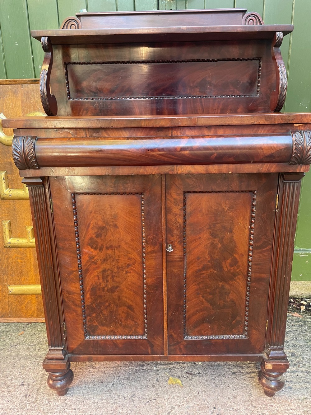 An early nineteenth-century George IV flame mahogany chiffonier featuring a raised shelf gallery with scroll supports above a two-door cabinet supported on turned bun feet. View II