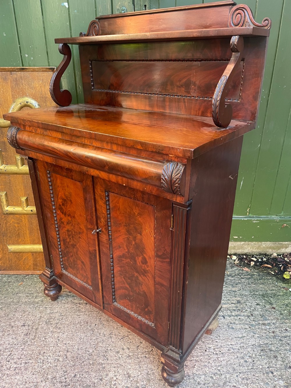 An early nineteenth-century George IV flame mahogany chiffonier featuring a raised shelf gallery with scroll supports above a two-door cabinet supported on turned bun feet. View IX