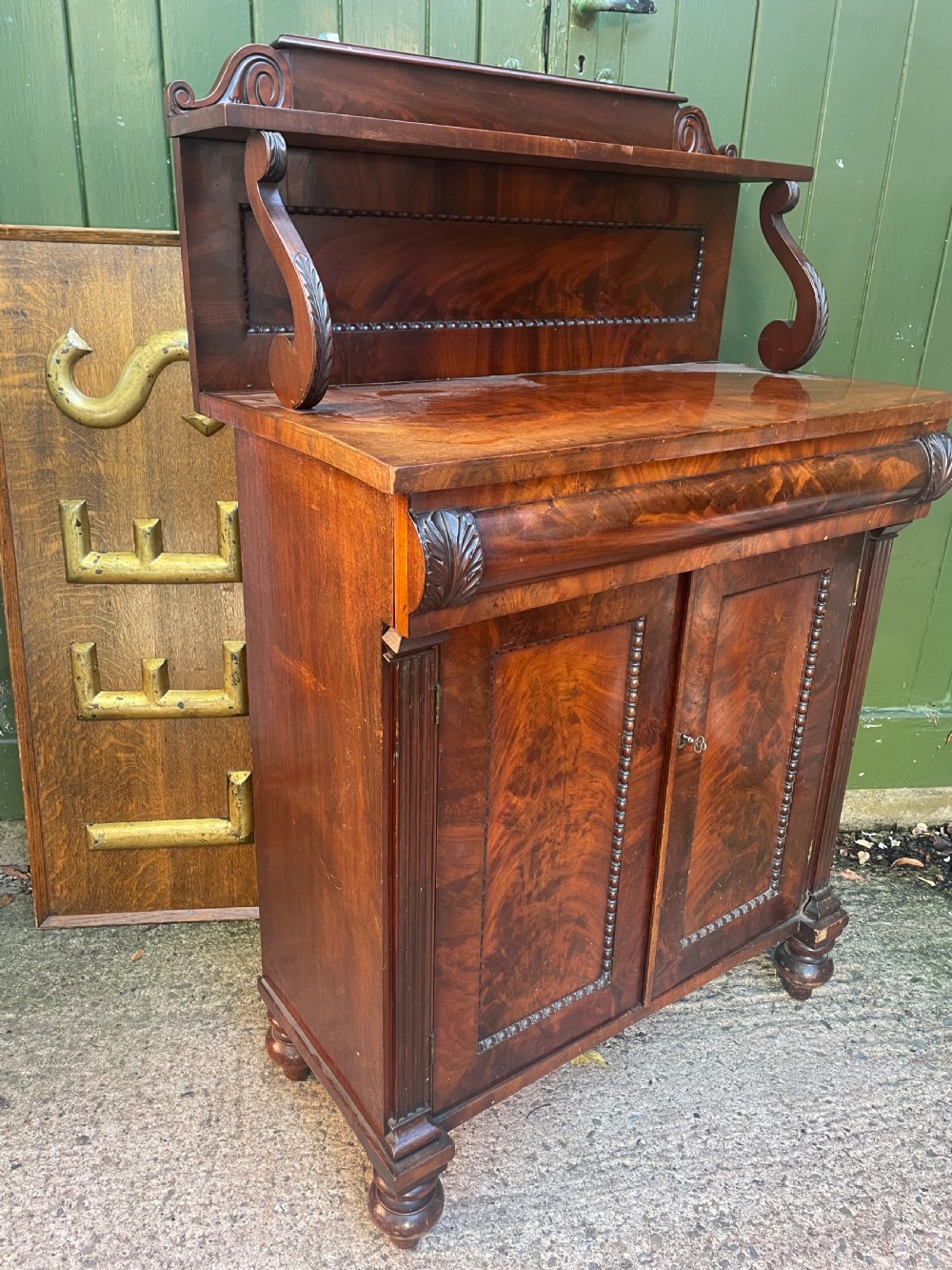 An early nineteenth-century George IV flame mahogany chiffonier featuring a raised shelf gallery with scroll supports above a two-door cabinet supported on turned bun feet. View X