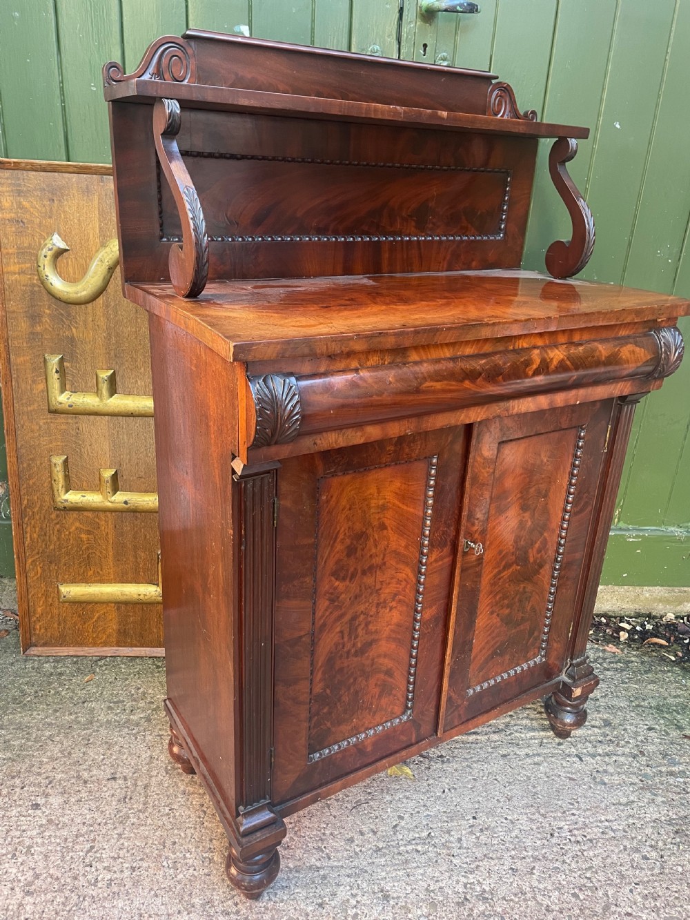 An early nineteenth-century George IV flame mahogany chiffonier featuring a raised shelf gallery with scroll supports above a two-door cabinet supported on turned bun feet. View XII